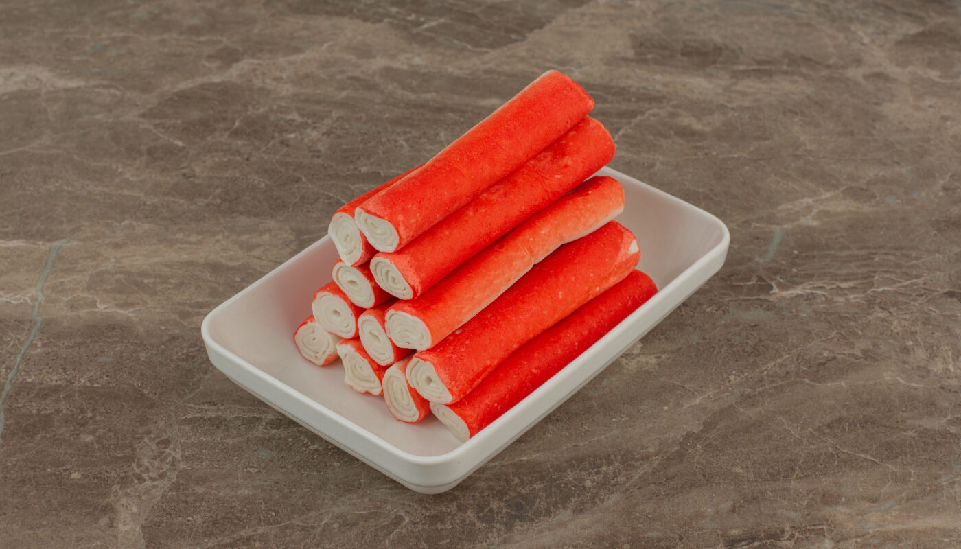 Крабовые палочки лежат на белом прямоугольном блюдце на сером столе